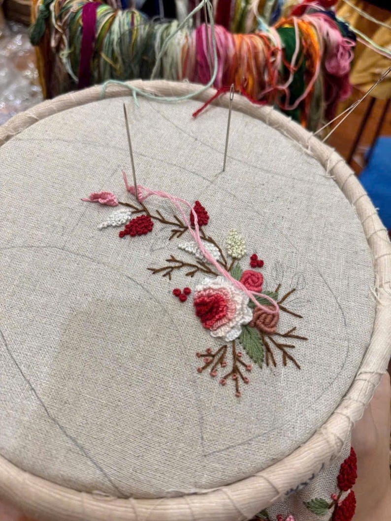 Puede incluir: Primer plano de un dise&ntilde;o de bordado floral en un trozo de tela blanca estirada en un aro de bordado de madera. El dise&ntilde;o presenta flores rosas y rojas con hojas verdes.