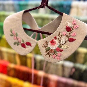 Puede incluir: Un par de cuellos de lino beige con delicados dise&ntilde;os florales bordados en rosa y rojo. Los cuellos est&aacute;n colgados en una percha de terciopelo.