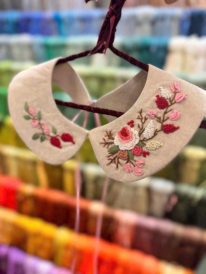Puede incluir: Un par de cuellos de lino beige con delicados dise&ntilde;os florales bordados en rosa y rojo. Los cuellos est&aacute;n colgados en una percha de terciopelo.