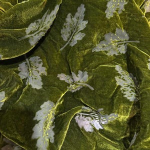 May include: A piece of olive green velvet fabric featuring a repeating pattern of light grey, leaf-shaped motifs. The fabric has a soft, plush texture with visible folds and drapes that create highlights and shadows across the surface.