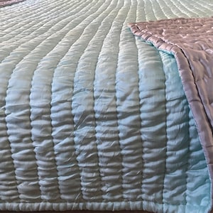 May include: A quilted bedspread featuring a light mint green top surface with vertical channel stitching and a folded corner revealing a smooth, solid lavender underside. The fabric has a subtle sheen and a soft, textured, rippled appearance.