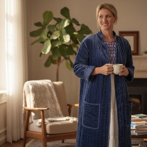 May include: A woman wearing a long, quilted navy blue robe with large pockets, holding a white mug. The robe is over a patterned top and white dress. The scene is a well-lit living room with a fireplace, chair, and a large plant.