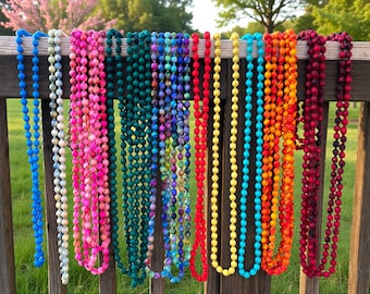 May include: A collection of beaded necklaces in various colors, including blue, pink, green, purple, red, yellow, turquoise, orange, and burgundy, hanging on a wooden fence. The necklaces are made of small, round beads.