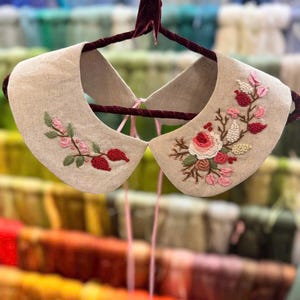 Puede incluir: Un cuello de lino beige con un delicado bordado floral en tonos rosa, rojo y verde. El cuello est&aacute; unido a una cinta rosa y est&aacute; colgado de una percha de terciopelo.