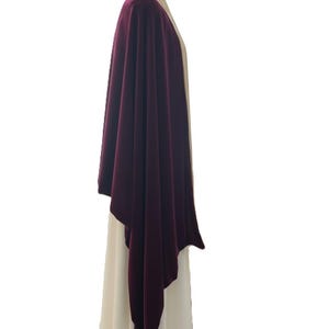 May include: A deep burgundy velvet wrap draped over a cream-colored garment. The wrap features a smooth, soft texture and an asymmetrical hemline that hangs down the side, contrasting with the light-colored base layer worn on a mannequin.