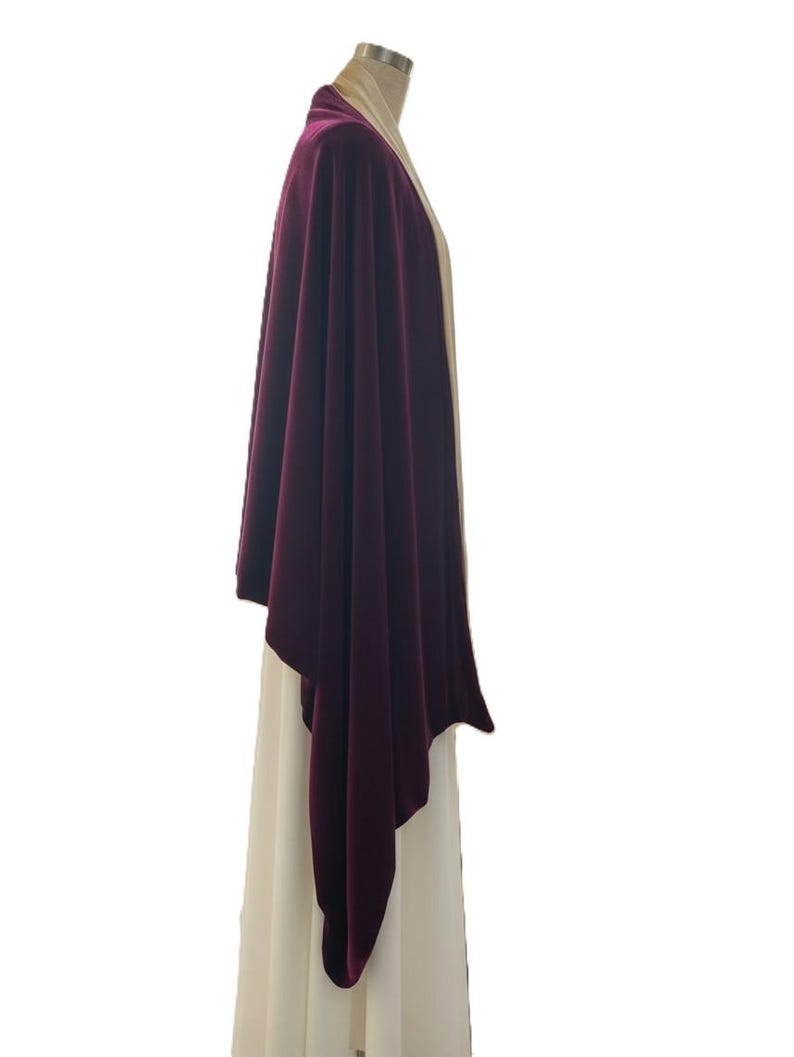 May include: A deep burgundy velvet wrap draped over a cream-colored garment. The wrap features a smooth, soft texture and an asymmetrical hemline that hangs down the side, contrasting with the light-colored base layer worn on a mannequin.