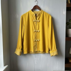 May include: A mustard yellow velvet shirt with a mandarin collar and long sleeves. The shirt features three sets of decorative frog closures down the front. The cuffs are folded back. The shirt is hanging on a wooden hanger.