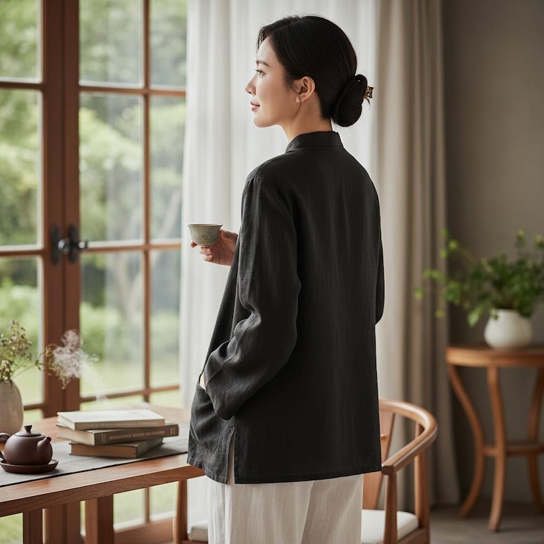 May include: A dark grey linen tunic with long sleeves and a mandarin collar. The tunic is worn with white trousers. A small ceramic cup is held. The scene is set near a window with natural light.