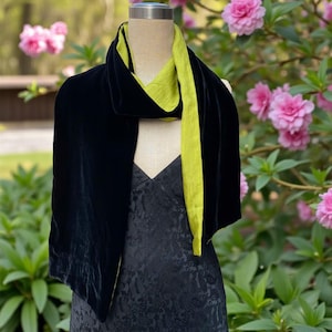 May include: A long scarf featuring a deep black velvet exterior and a contrasting lime green interior lining, draped over a mannequin wearing a black dress with a subtle tonal pattern. The scarf displays a soft texture and a smooth, draped silhouette.