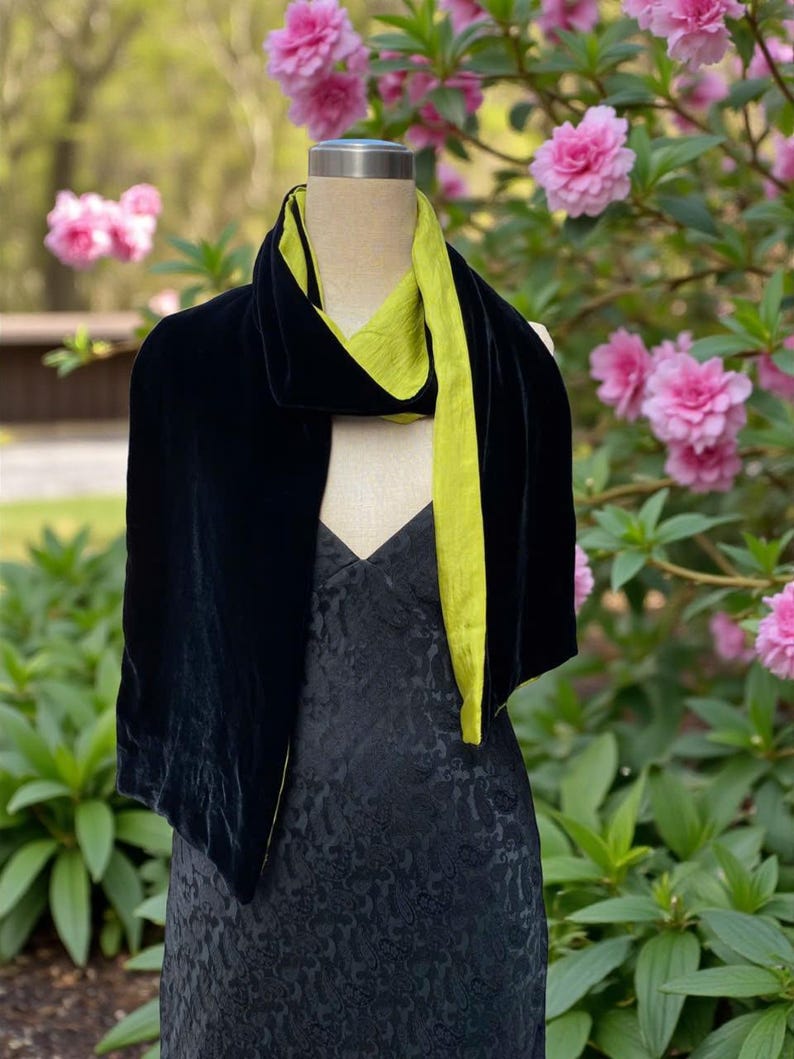 May include: A long scarf featuring a deep black velvet exterior and a contrasting lime green interior lining, draped over a mannequin wearing a black dress with a subtle tonal pattern. The scarf displays a soft texture and a smooth, draped silhouette.