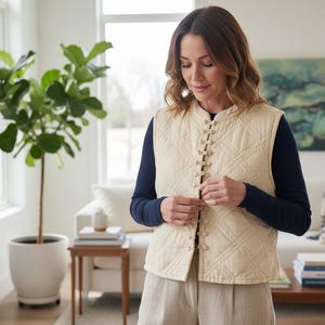 May include: A cream-coloured quilted vest with a diamond pattern and decorative toggles. The vest is worn over a navy blue long-sleeved shirt and paired with beige trousers. The scene is set in a well-lit living room with a large potted plant.