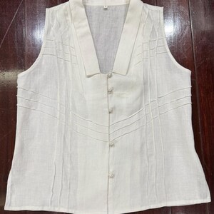 Puede incluir: Una blusa sin mangas de lino blanco roto con cuello en V y cierre de botones. Los paneles frontales tienen detalles cosidos. La tela es ligera, adecuada para climas c&aacute;lidos. La blusa es de color neutro.
