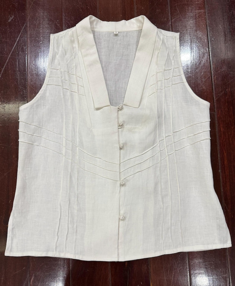 Puede incluir: Una blusa sin mangas de lino blanco roto con cuello en V y cierre de botones. Los paneles frontales tienen detalles cosidos. La tela es ligera, adecuada para climas c&aacute;lidos. La blusa es de color neutro.