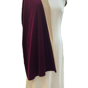 May include: A long, deep burgundy velvet shawl draped over a solid white sleeveless dress. The shawl features a smooth, rich texture and a contrasting beige lining visible along the folded edge, creating a layered look over the plain white gown.