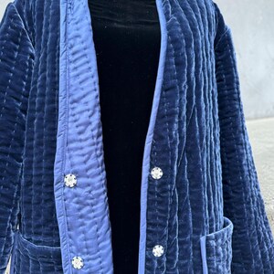 May include: A quilted navy blue velvet jacket with a contrasting light blue trim. The jacket features three decorative buttons and two pockets. The jacket is displayed on a mannequin.