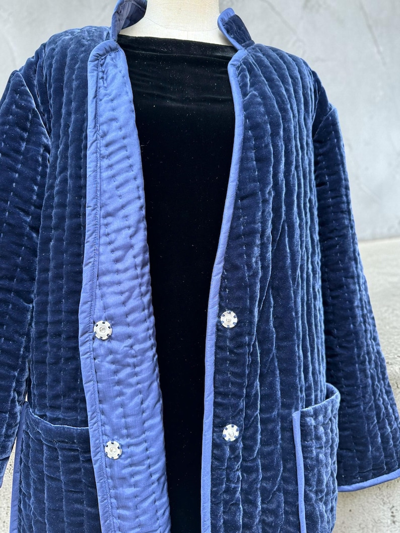May include: A quilted navy blue velvet jacket with a contrasting light blue trim. The jacket features three decorative buttons and two pockets. The jacket is displayed on a mannequin.