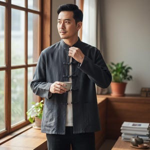 May include: A dark grey jacket with a mandarin collar and frog fastenings. The jacket features two front pockets and is worn over a white shirt. The person is holding a small ceramic cup. The jacket is a traditional style.