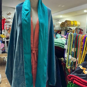 May include: A teal velvet wrap featuring a wide, smooth teal satin collar draped over a coral-colored sleeveless garment. The wrap has a soft, reflective texture and hangs loosely from the shoulders, showcasing a layered, open-front design.