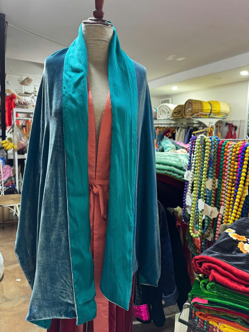 May include: A teal velvet wrap featuring a wide, smooth teal satin collar draped over a coral-colored sleeveless garment. The wrap has a soft, reflective texture and hangs loosely from the shoulders, showcasing a layered, open-front design.