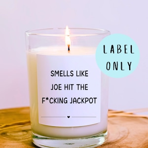 May include: A burning candle in a clear glass jar with a white label. The label says "Smells like Joe hit the f*cking jackpot." A light blue circle with the words "Label Only" is on the right. The candle is on a wooden surface.