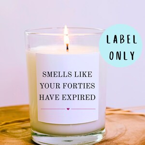 May include: A burning candle in a clear glass jar with a white label. The label says "Smells Like Your Forties Have Expired" in black text, with a small pink heart. A light blue circle with the words "Label Only" is in the background.