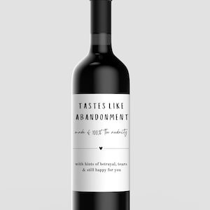 May include: A black wine bottle with a white label. The label reads "TASTES LIKE ABANDONMENT" and "made of 100% the audacity" with the text "with hints of betrayal, tears & still happy for you".