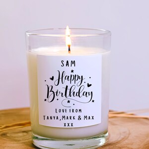 May include: A lit candle in a clear glass jar. The candle has a white label with the words "SAM Happy Birthday" in black script. The label also includes the text "LOVE FROM TANYA, MARK & MAX" and "XXX". The candle is on a wooden surface.