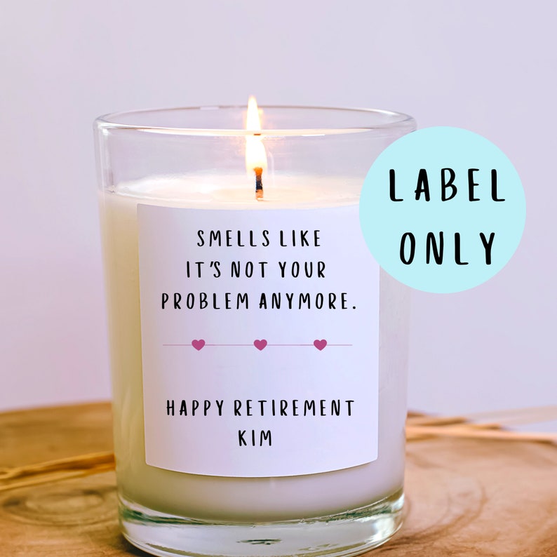 May include: White candle with a label that reads "Smells like it's not your problem anymore. Happy Retirement Kim" with three pink hearts. The candle is lit and the label is on a clear glass jar.