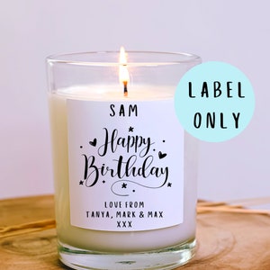 May include: A lit candle in a clear glass jar with a white label. The label reads "SAM Happy Birthday" with additional text "LOVE FROM TANYA, MARK & MAX XXX". The candle is on a wooden surface.