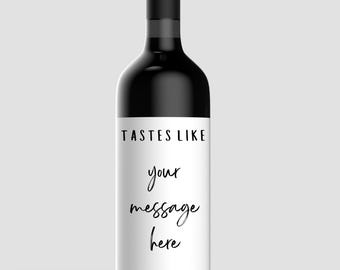 以下が含まれることがあります： 白いラベルが付いた黒いワインボトル。ラベルには上部に「TASTES LIKE」、下に筆記体で「your message here」と書かれています。ボトルには黒いキャップが付いています。