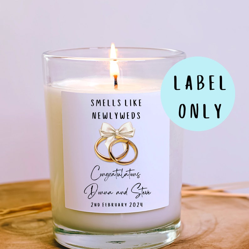 Puede incluir: Vela blanca con una etiqueta que dice "Huele a reci&eacute;n casados" y tiene un dise&ntilde;o de anillos dorados con un lazo blanco. La etiqueta tambi&eacute;n dice "Felicidades Donna y Steve 2 de febrero de 2024".