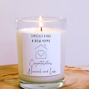 May include: A white candle in a clear glass jar with a label that reads "Smells like a new home" and a house with a heart inside. The label also says "Congratulations Hannah and Luke".
