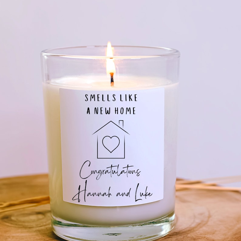 May include: A white candle in a clear glass jar with a label that reads "Smells like a new home" and a house with a heart inside. The label also says "Congratulations Hannah and Luke".