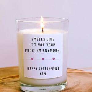 May include: A white candle with a black label that reads "Smells like it's not your problem anymore. Happy Retirement Kim" with three pink hearts. The candle is lit and in a clear glass jar.
