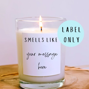 May include: A clear glass candle jar with a white label that says "Smells Like" and has a space for a custom message. The candle is lit and the label has a light blue circle with the text "LABEL ONLY" on it.