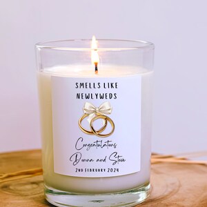 Puede incluir: Una vela blanca con una etiqueta que dice "Smells Like Newlyweds". La etiqueta tambi&eacute;n presenta un lazo blanco con dos anillos dorados y el texto "Congratulations Donna and Steve 2nd February 2024".