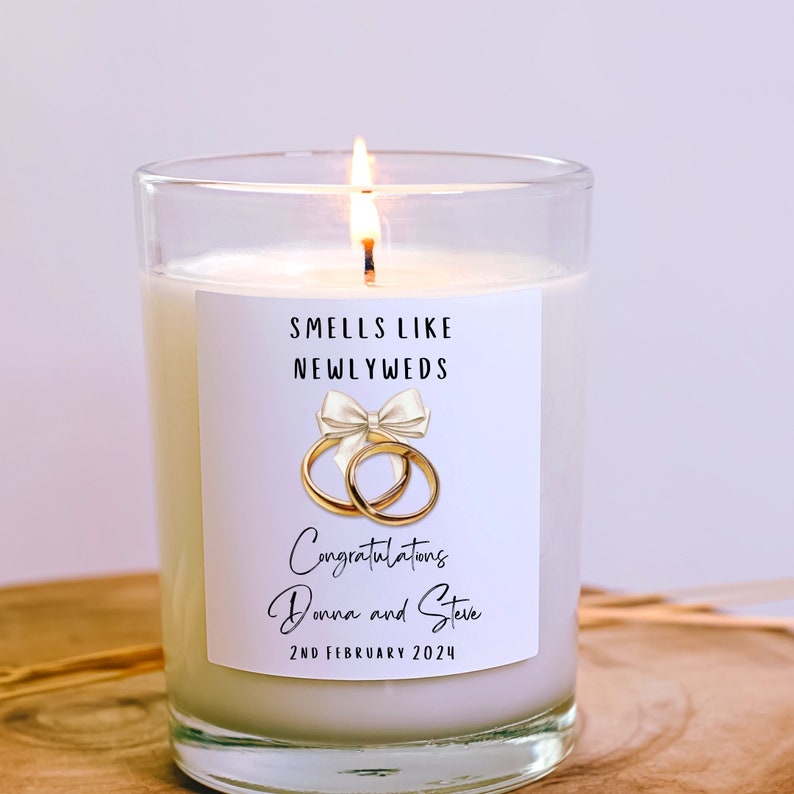Puede incluir: Una vela blanca con una etiqueta que dice "Smells Like Newlyweds". La etiqueta tambi&eacute;n presenta un lazo blanco con dos anillos dorados y el texto "Congratulations Donna and Steve 2nd February 2024".