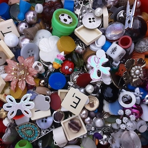 May include: A close-up of a mixed collection of craft supplies, including buttons, beads, and decorative items. The image shows a green football-shaped item, flower-shaped pieces, and Scrabble tiles. The colour palette includes pink, white, blue, and gold.