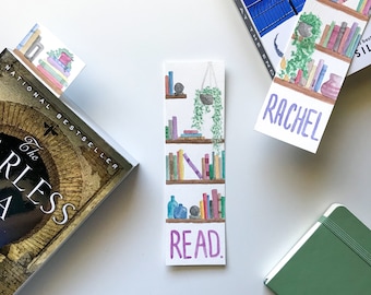 Peut inclure: Un marque-page aquarelle représentant une bibliothèque avec des livres, une plante suspendue et le mot "READ". Le marque-page est blanc avec du texte violet et se trouve sur une surface blanche.