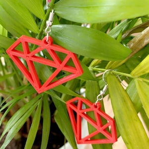 May include: A pair of red geometric diamond-shaped earrings with silver hooks. The earrings are hanging from a green plant.