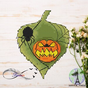 May include: A cross-stitch design of a green leaf with a carved orange pumpkin and a black spider. The leaf has detailed veins. The design is set against a white wooden background. A Halloween-themed craft project.