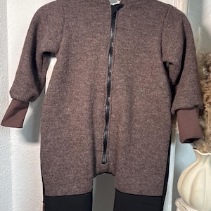 Könnte beinhalten: Brauner Kapuzen-Overall mit durchgehendem schwarzem Reißverschluss und schwarzen Kniepatches. Die Ärmel und Knöchel haben braune Bündchen. Der Overall hängt an einer weißen Holzleiter.