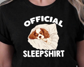 Puede incluir: Camiseta negra con texto blanco que dice "OFFICIAL SLEEP SHIRT". Una imagen de dibujos animados de un Cavalier King Charles Spaniel durmiendo en una almohada blanca está por encima del texto.