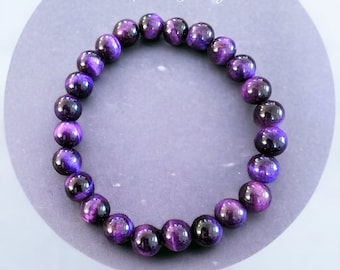 May include: Purple Tiger Eye gemstone bracelet with round beads. The bracelet is on a gray background.
