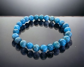 Puede incluir: Un brazalete de piedra de apatita azul con cuentas redondas. El brazalete está sobre una superficie reflectante y el texto "Blue Apatite" y "Bubbs Bracelets" es visible.