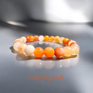 May include: A beaded bracelet featuring a variety of orange, yellow, and white gemstones. The bracelet is made of round beads in varying shades, creating a gradient effect. The bracelet is displayed on a gray surface.