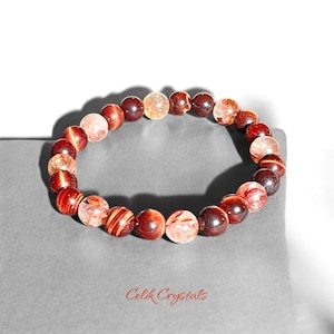 May include: A red and gold beaded bracelet with a mix of round and faceted beads. The bracelet is on a gray surface with the text "Celik Crystals" printed below.