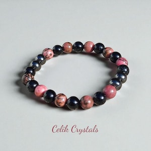 Puede incluir: Un brazalete de cuentas con cuentas alternas de piedras preciosas negras y rosas. Las cuentas rosas tienen un aspecto moteado, mientras que las cuentas negras son lisas y pulidas. El texto "Celik Crystals" está en la parte inferior.