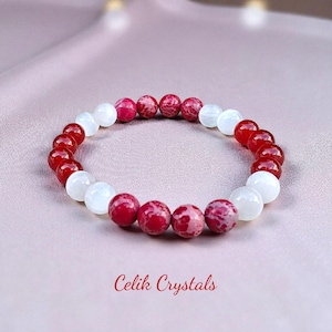 May include: A beaded bracelet featuring alternating red and white round beads. The red beads have a speckled pattern. The bracelet is displayed on a pink satin surface. The text "Celik Crystals" is at the bottom.