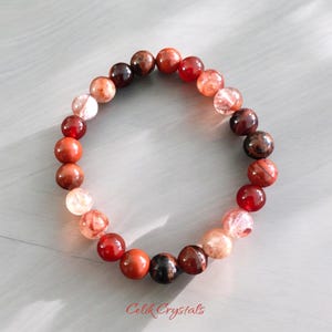 May include: A red, orange, and brown gemstone bracelet with a gradient of color. The bracelet is made of round beads and is approximately 7 inches in length.